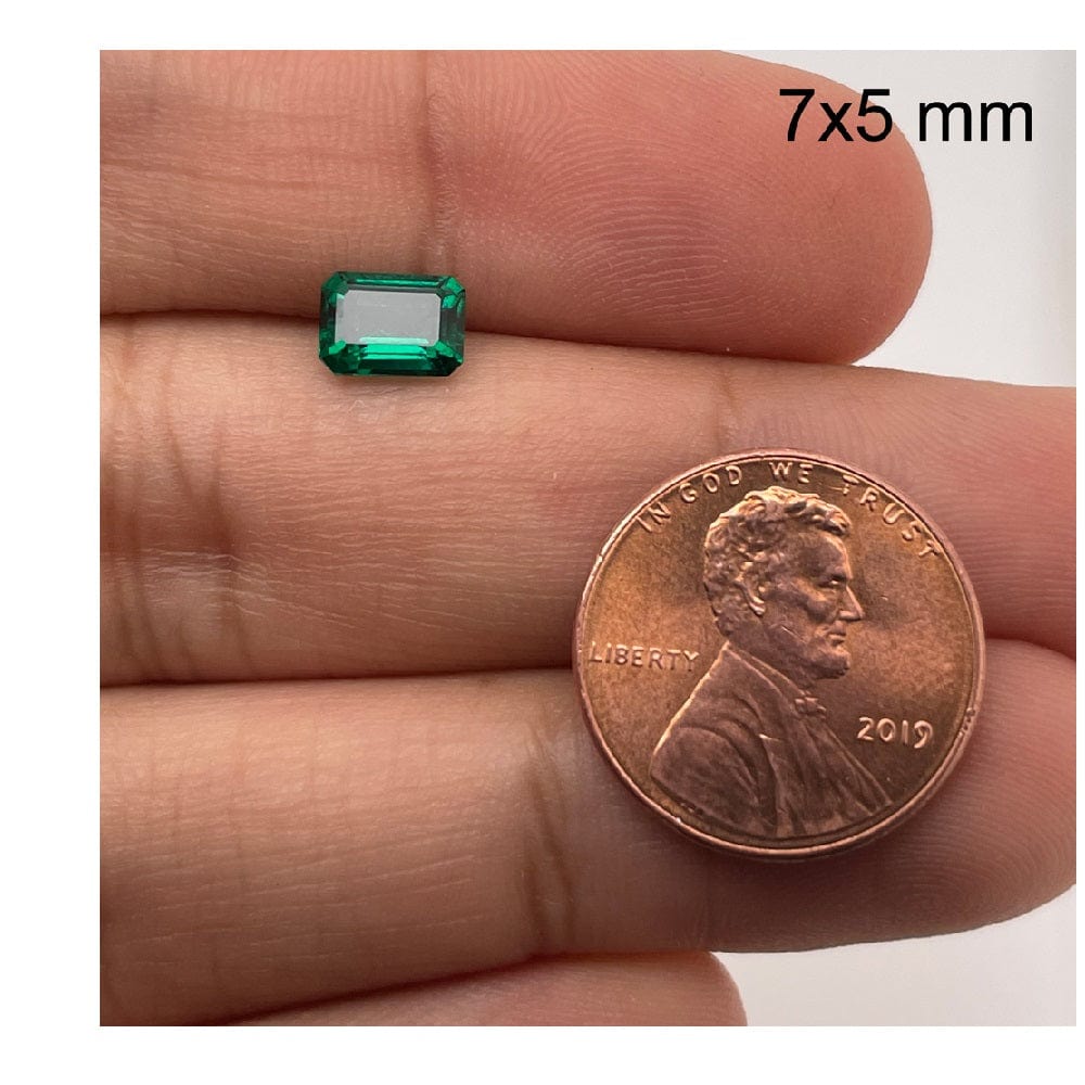 A hand displays a 7x5 mm 7x5 Exceptional Emerald Color Lab Created Emerald beside a 2019 U.S. penny for size comparison, with 7x5 mm text in the top right corner.