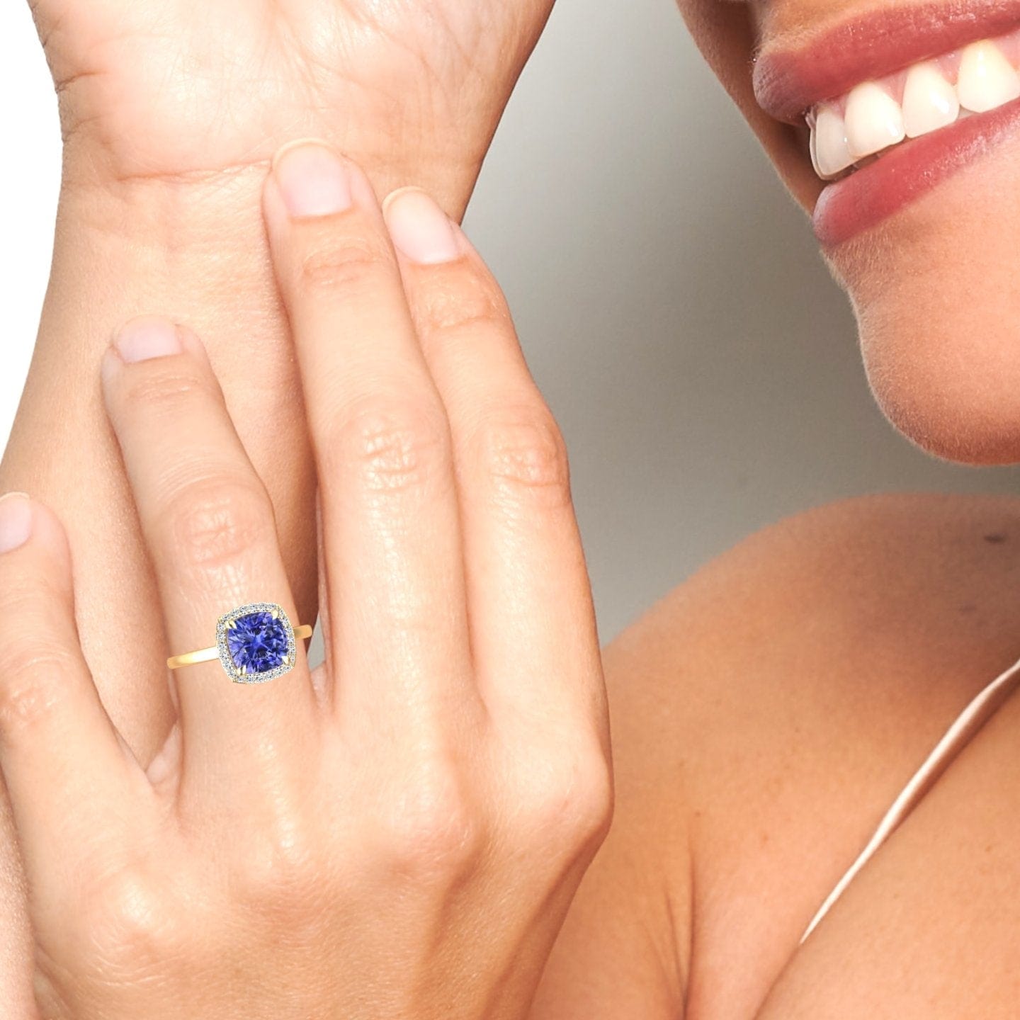 A close-up of a smiling person’s hand highlights the Ziva Tanzanite & Diamond Halo Ring, featuring a large square blue gemstone surrounded by sparkling diamonds. The person has light skin and wears a thin strap top.