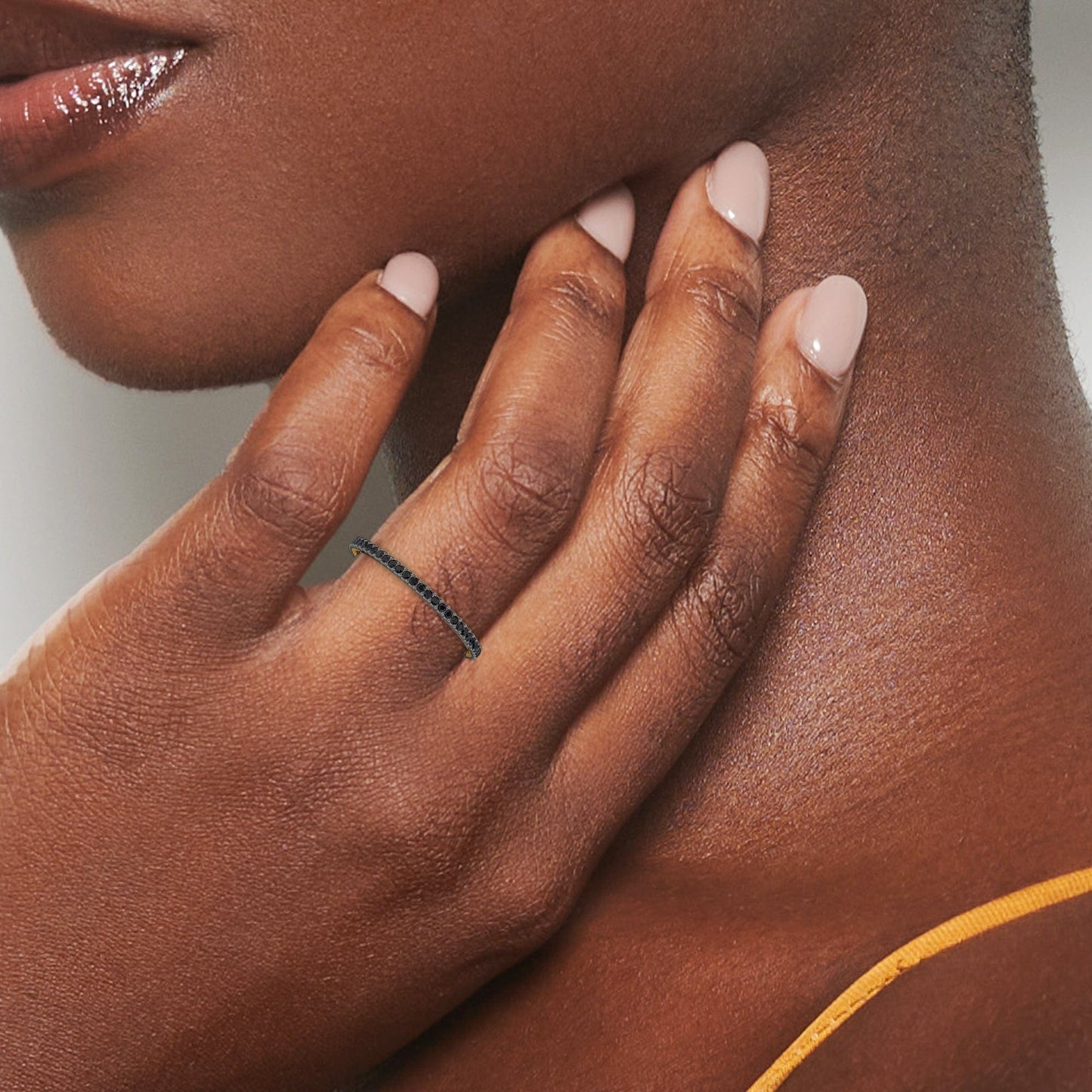 A womanโs nude polished nails gently touch her neck as she wears the Black Diamond Ballad Eternity Ring - Colleen, a delicate thin black band. A yellow strap appears on her shoulder.