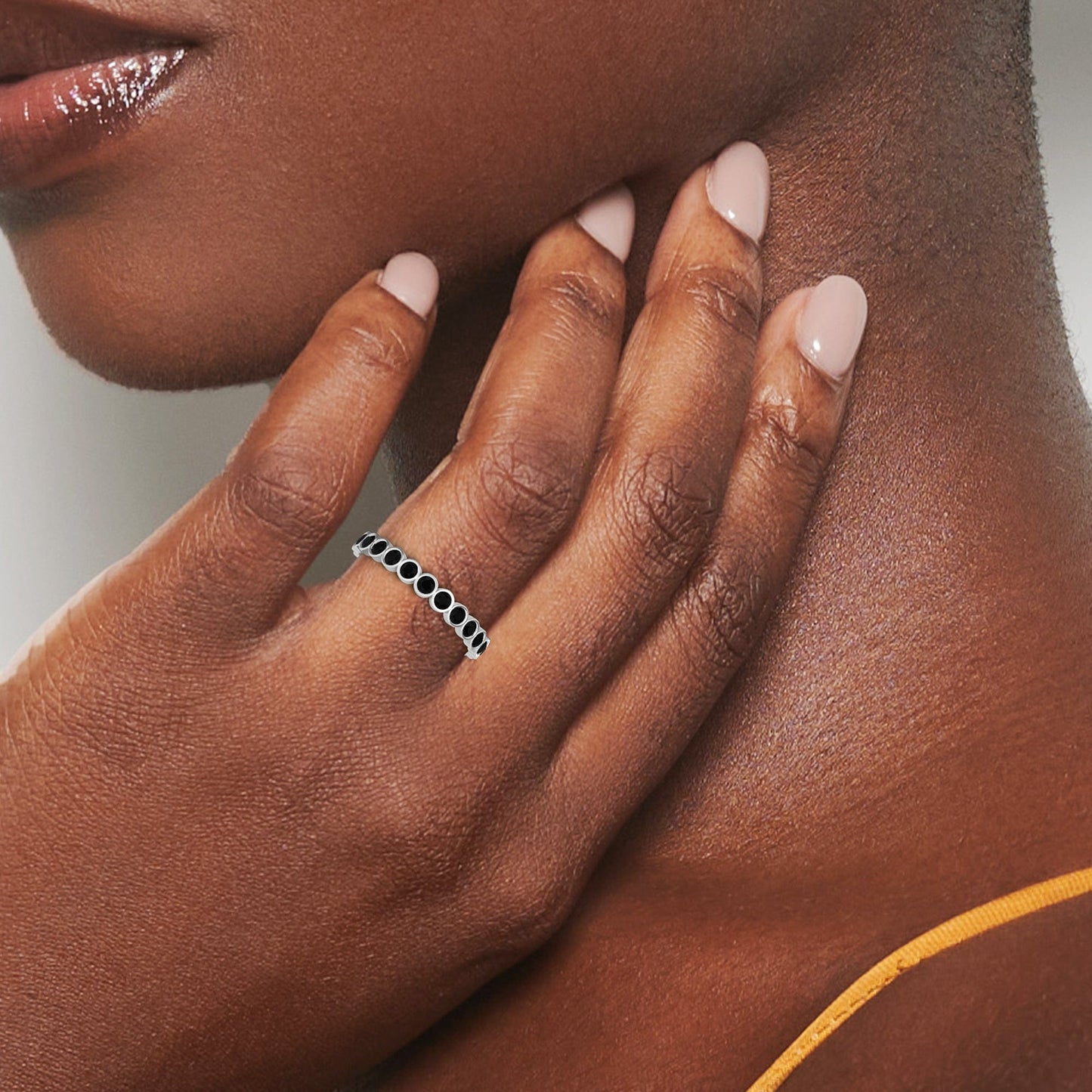 A close-up of a hand with light pink manicured nails touching the neck, wearing the Zara Black Diamond Bezel Set Eternity Band Ring. The person is in a yellow spaghetti strap top.
