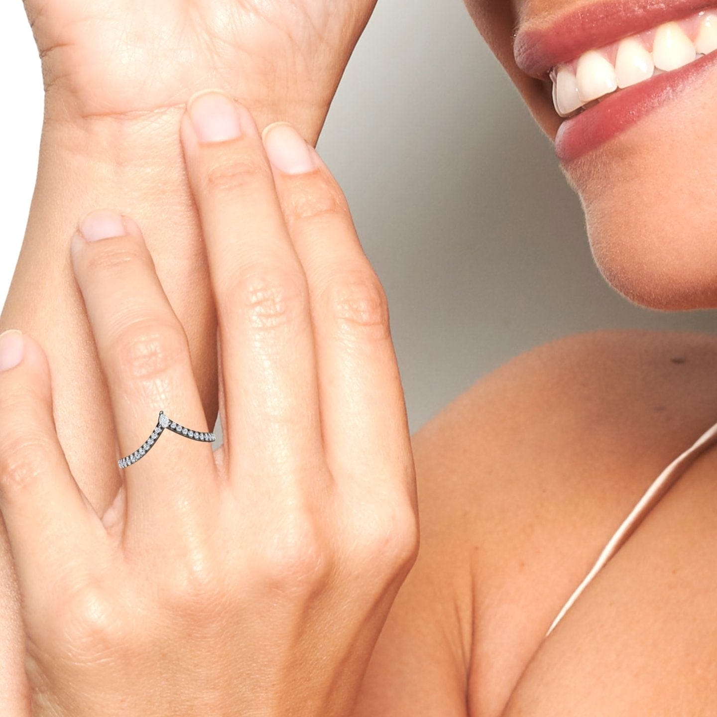 A close-up of a smiling personโs hand wearing the White Diamond Classic Chevron Wedding Band - Mei-Lin, a delicate V-shaped silver ring with small stones. Their face is partially visible, showing white teeth and smooth skin.