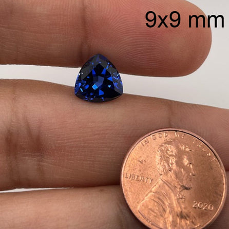 A hand holds a 9mm Exceptional Pear Trillion Created Blue Sapphire next to a U.S. penny for size reference. The text 9x9 mm appears at the top right of the image.