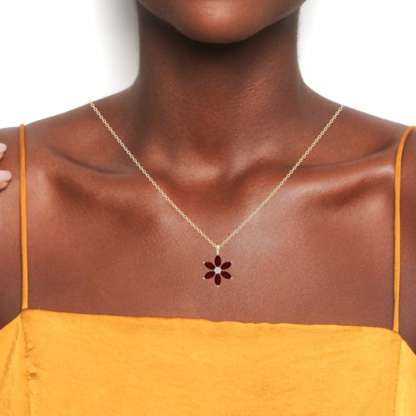 A person wears the Lab Grown Ruby & Diamond Floral Necklace - Erin, featuring a thin gold chain with a red flower-shaped pendant, paired with a yellow spaghetti strap top against a white background.