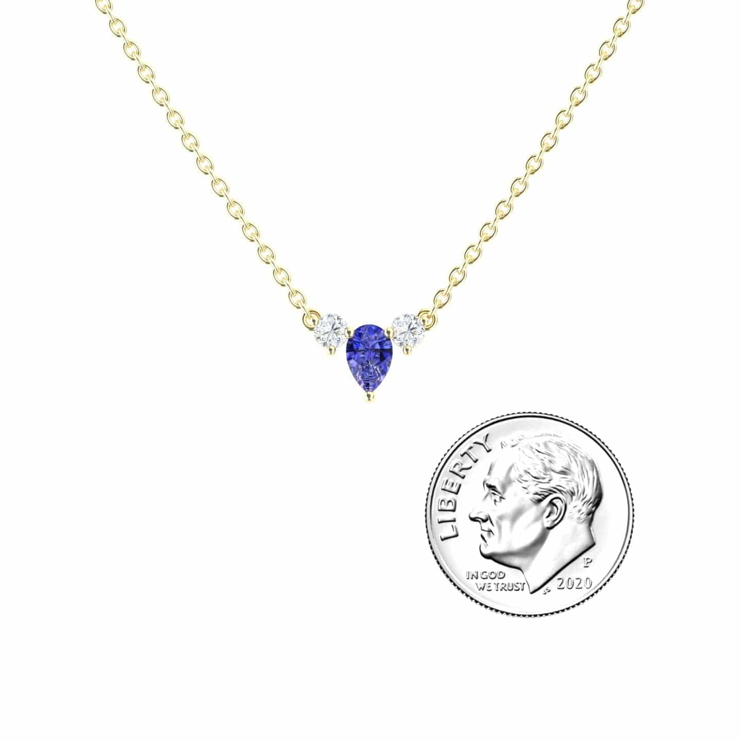 The Tanzanite & Diamond Three-Stone Necklace - Jina features a teardrop-shaped blue tanzanite pendant with two small diamond accents on a gold chain, shown beside a U.S. dime for size reference.