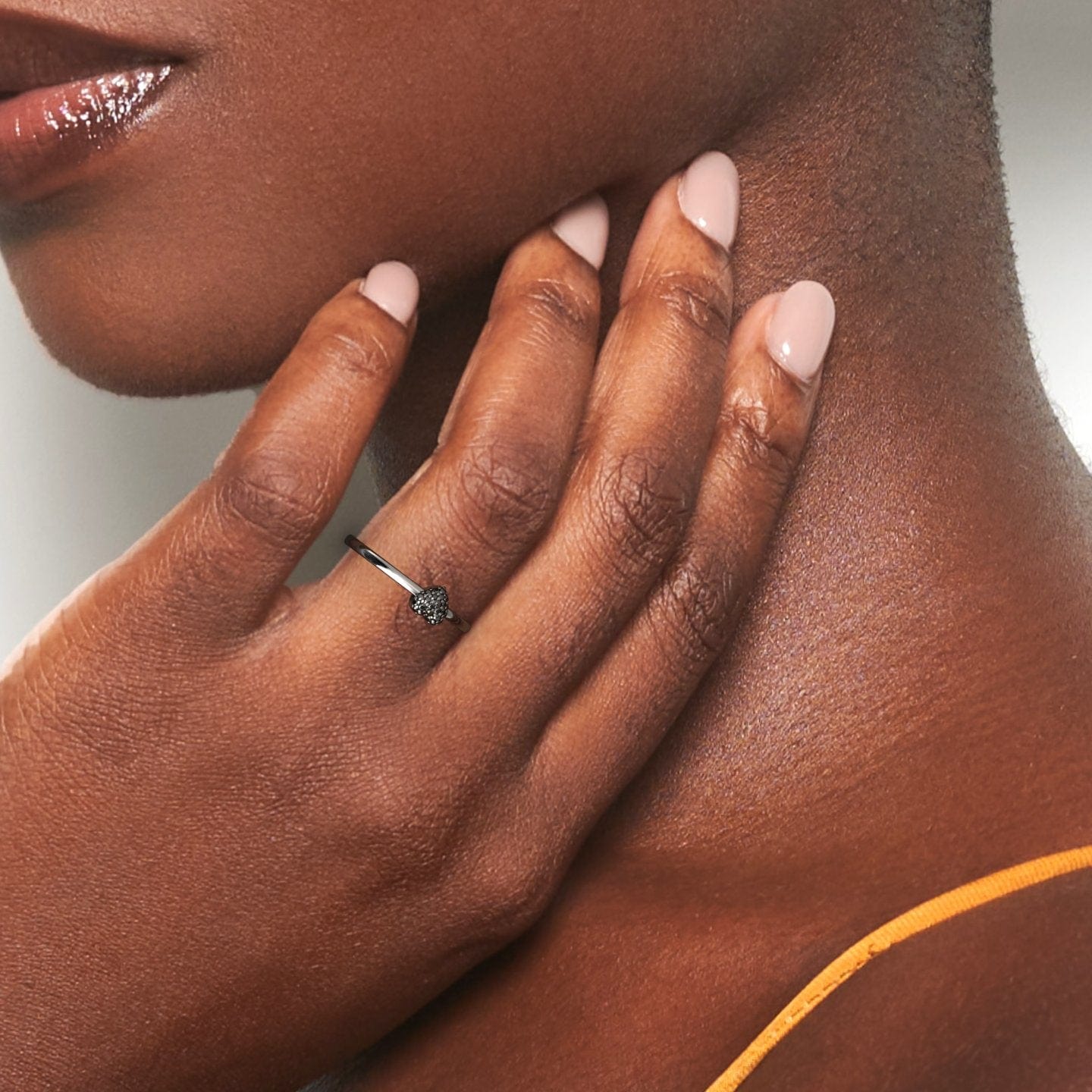 A close-up of a woman’s hand with light pink nails gently touching her neck. She has dark skin, wears a yellow-orange strap, and is wearing the Bonita Black Diamond Pave Heart Ring—a delicate black ring with a small decorative heart detail.